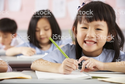 Young Female Student Working At Desks In Chinese School Classroom Smiling To Camera